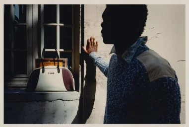 Man wearing a textured blue sweater standing beside a white building window with a duffel bag on the sill