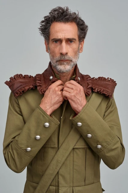 Mature man in green military-style jacket with a brown ruffled collar adjusts his collar, in a medium studio shot against a grey backdrop.