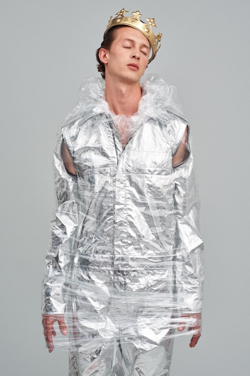 Fashion shot of a man wearing a gold crown and wrapped in clear plastic, standing in a photo studio with eyes closed.