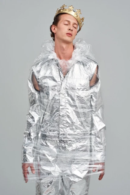 Fashion shot of a man wearing a gold crown and wrapped in clear plastic, standing in a photo studio with eyes closed.