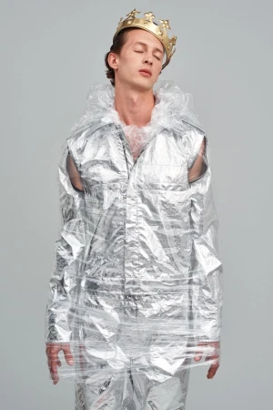 Fashion shot of a man wearing a gold crown and wrapped in clear plastic, standing in a photo studio with eyes closed.