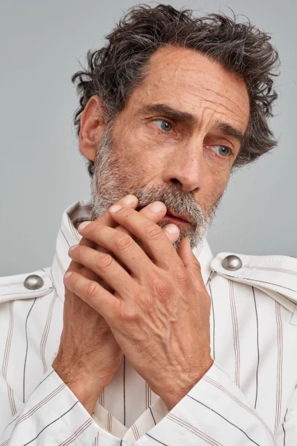 Portrait of a middle-aged man with grey hair and beard wearing a striped shirt, hands clasped, looking off to the side in a photo studio.