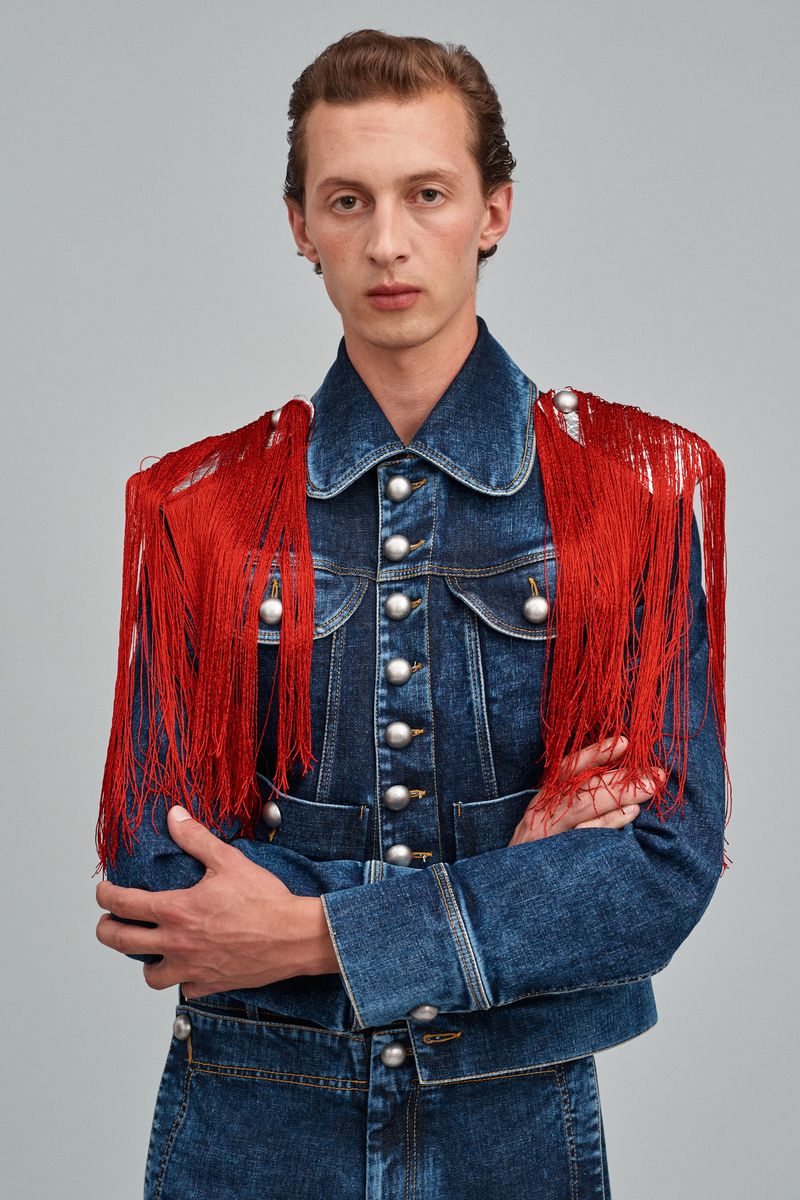 Adult male poses indoors wearing a blue denim jacket adorned with red fringe and silver buttons, against a neutral background.