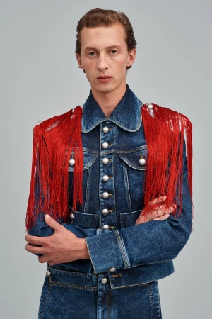 Adult male poses indoors wearing a blue denim jacket adorned with red fringe and silver buttons, against a neutral background.