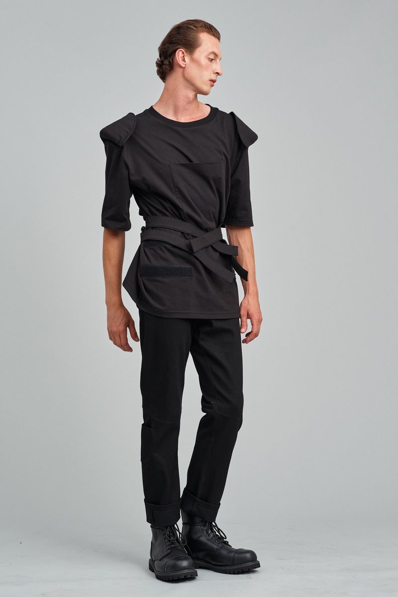 A slim adult male model in a black outfit is standing in a full shot against a gray background in a studio setting for a fashion shoot.