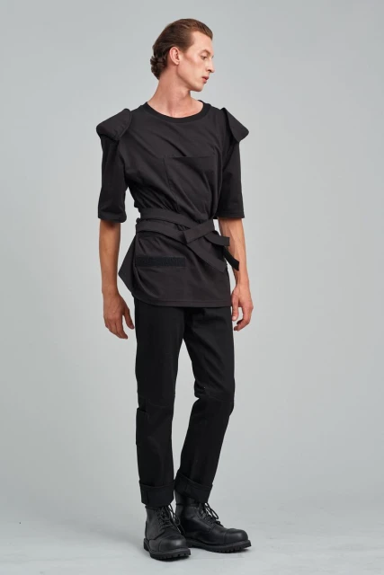 A slim adult male model in a black outfit is standing in a full shot against a gray background in a studio setting for a fashion shoot.