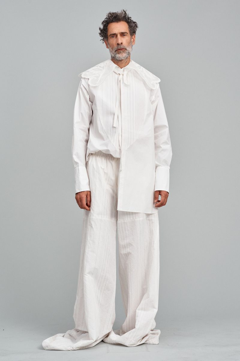Full-length portrait of an adult male with curly hair, wearing a white, oversized shirt with a large collar and wide-leg pants, in a studio.
