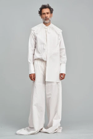 Full-length portrait of an adult male with curly hair, wearing a white, oversized shirt with a large collar and wide-leg pants, in a studio.