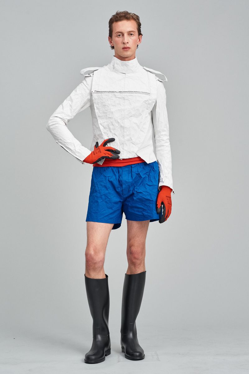Full-length studio shot of a young adult man in a white jacket, blue shorts, orange gloves, and black boots posing against a gray backdrop.