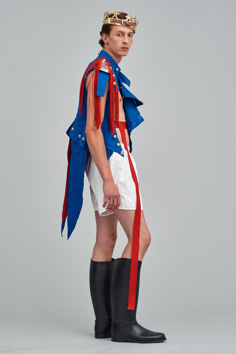 Side view of a fashion model wearing a blue vest with red fringe, white shorts with red stripes, black boots, and a gold crown.
