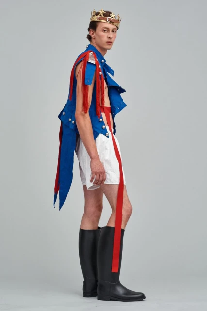 Side view of a fashion model wearing a blue vest with red fringe, white shorts with red stripes, black boots, and a gold crown.