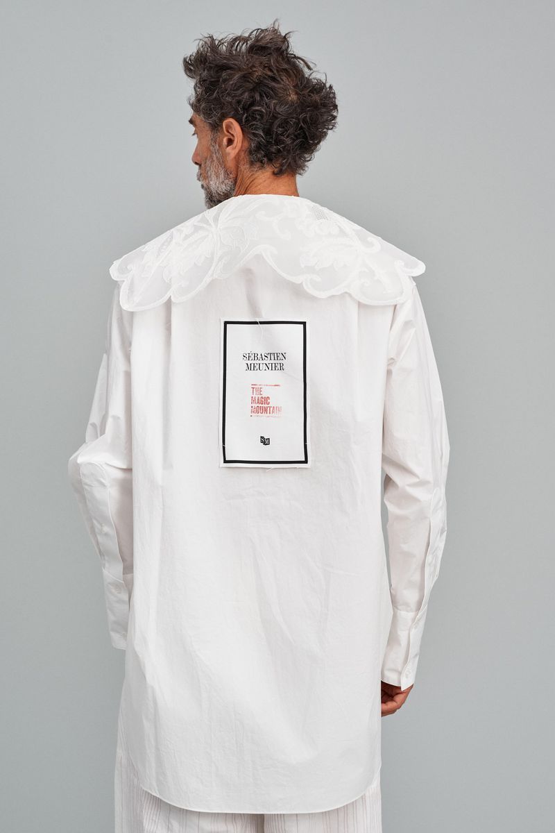 Adult male stands in photo studio wearing a white shirt with a large ruffled collar and black and red text design on the back.