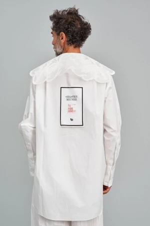 Adult male stands in photo studio wearing a white shirt with a large ruffled collar and black and red text design on the back.