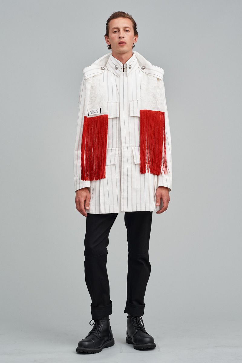 Male fashion model in striped jacket with red fringe posing in studio with black pants and boots, showcasing avant-garde style.