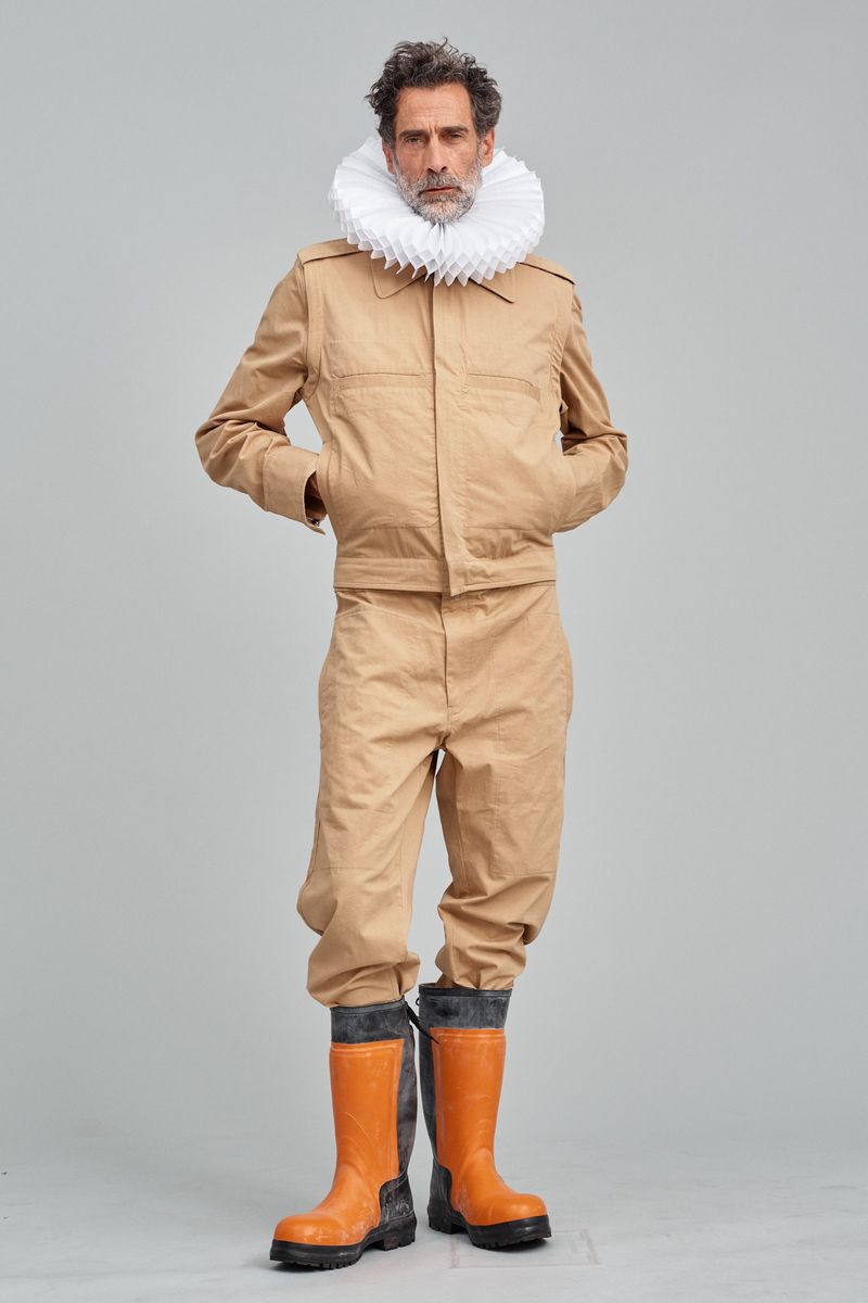 Mature male model in tan utility suit and orange rubber boots with white neck ruff standing in photo studio against grey background.