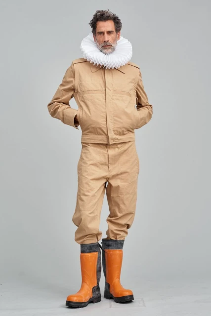 Mature male model in tan utility suit and orange rubber boots with white neck ruff standing in photo studio against grey background.