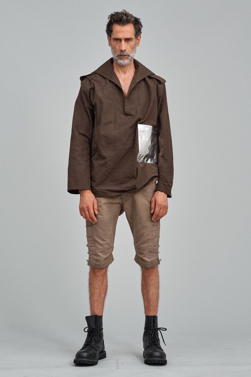 Full-length portrait of a mature man in a brown top and shorts, standing against a neutral gray backdrop, with black boots.