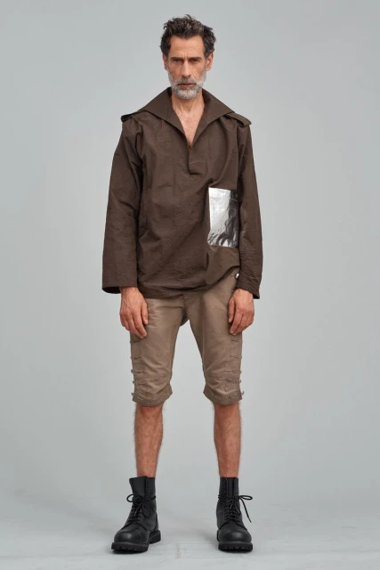 Full-length portrait of a mature man in a brown top and shorts, standing against a neutral gray backdrop, with black boots.