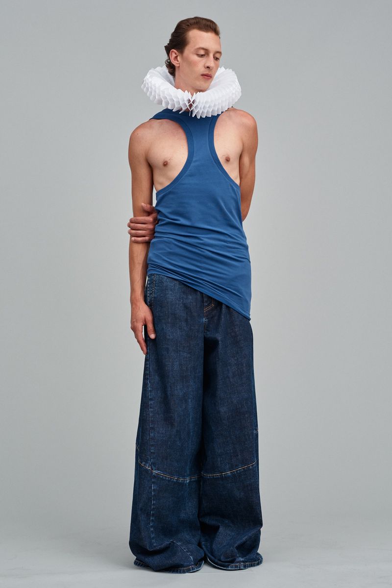 Full-length studio shot of adult male in blue tank top and wide-legged denim pants, featuring a pleated white neck ruff.
