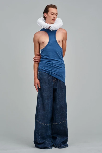 Full-length studio shot of adult male in blue tank top and wide-legged denim pants, featuring a pleated white neck ruff.