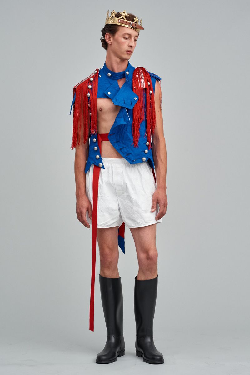 Man posing in a blue vest with red fringe, white shorts, black boots and a crown, standing in a full shot on a grey background.