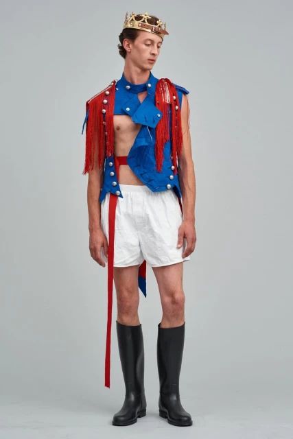 Man posing in a blue vest with red fringe, white shorts, black boots and a crown, standing in a full shot on a grey background.