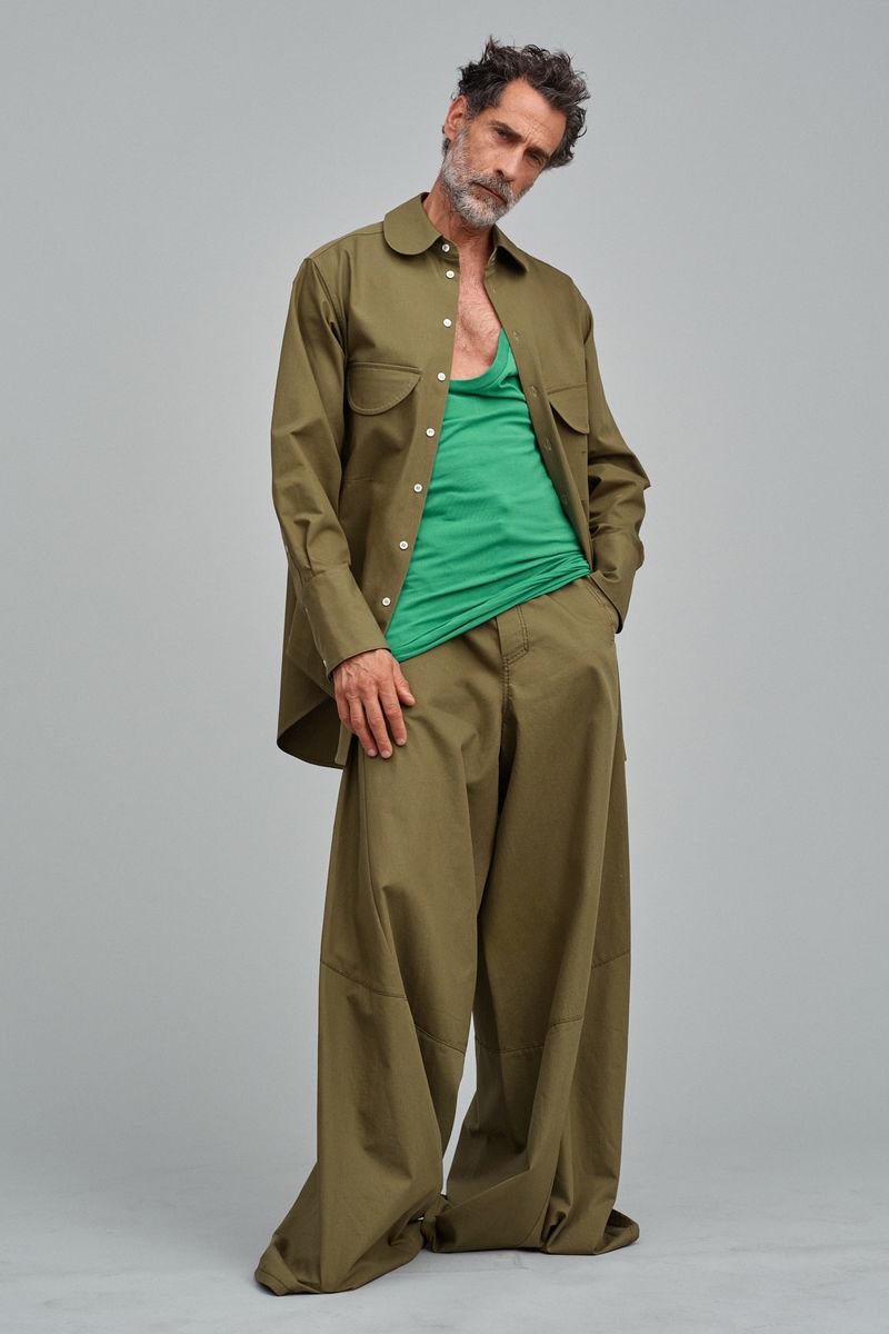 An adult male poses in a studio with loose green shirt, baggy green pants and matching green button-down shirt open. Clean fashion shot.