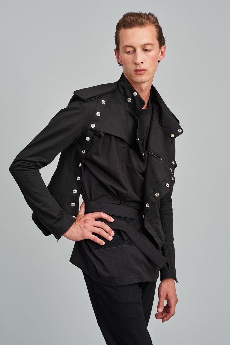 A slender man in a black military-style belted jacket looks away in a fashion shot against a grey backdrop in a studio setting.