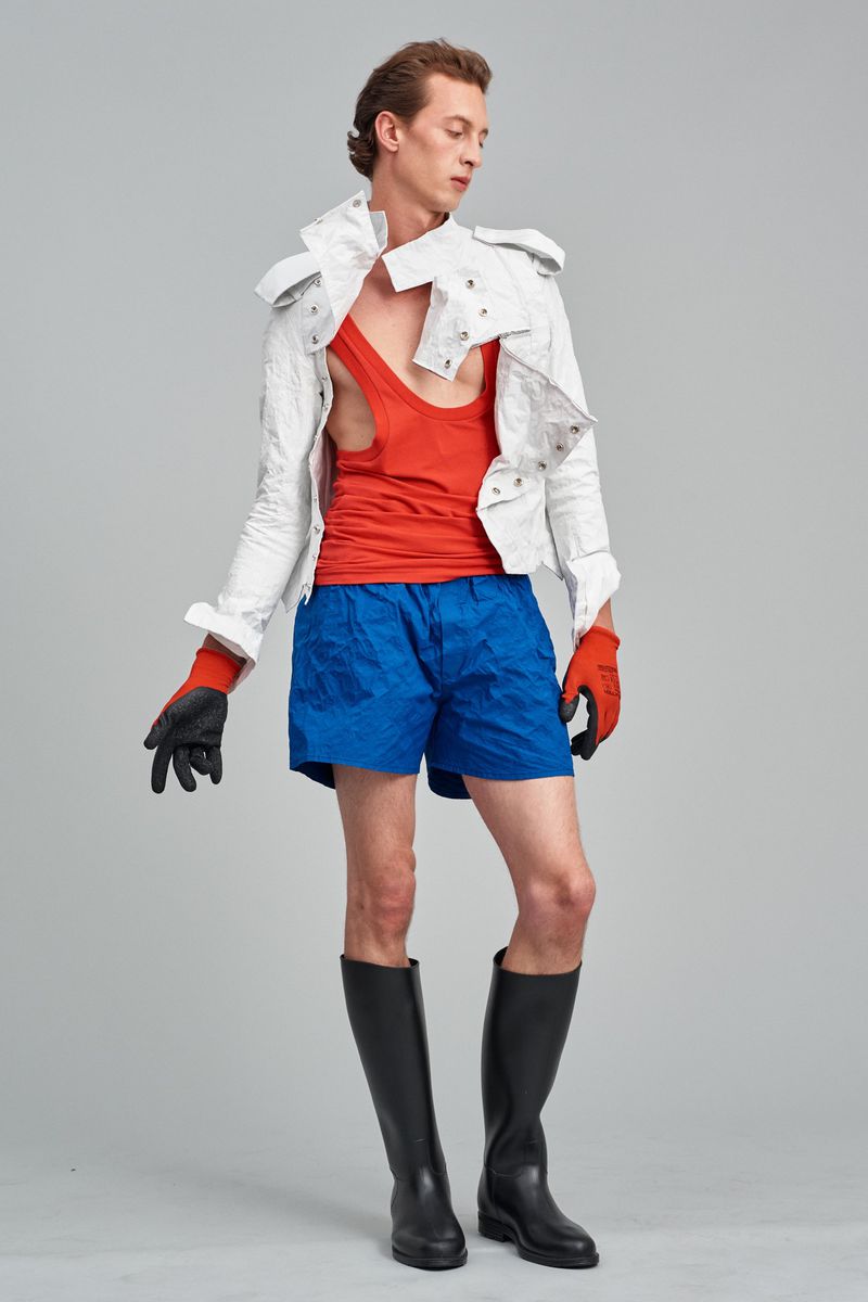 Male fashion model posing in a white jacket, orange tank top, blue shorts, black gloves, and boots in a light studio.