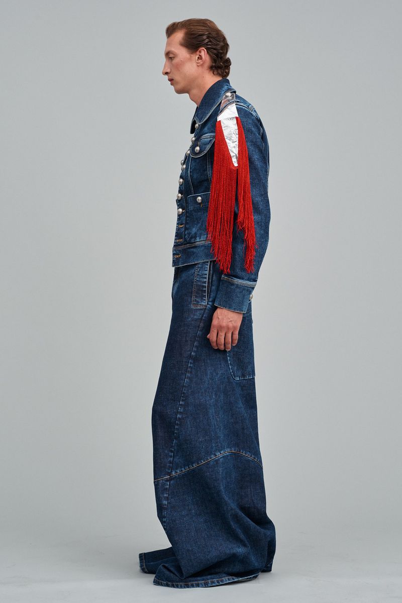 A slender man in a profile view is showcased in a blue denim jacket with red fringed epaulettes and baggy blue denim skirt-pants against grey.