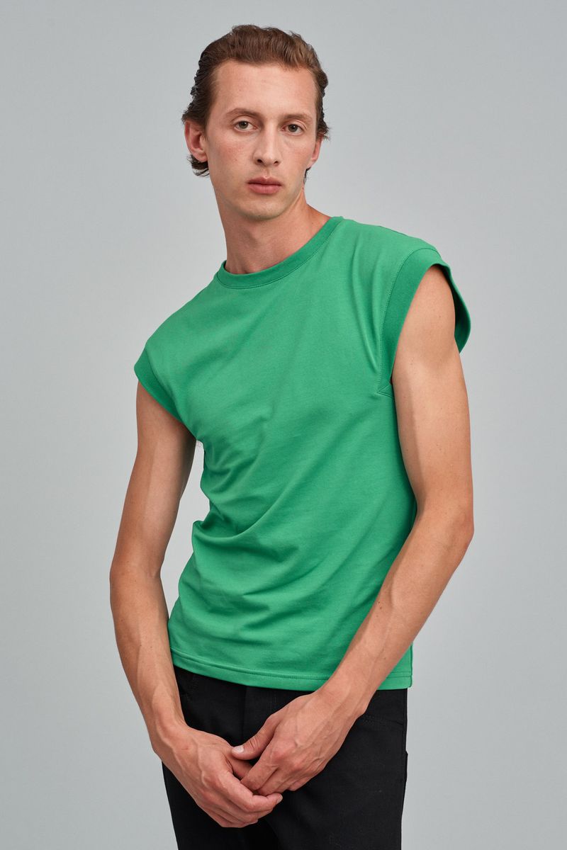 A fair-skinned adult male poses indoors wearing a green short-sleeved shirt and black pants, shot against a light gray backdrop.