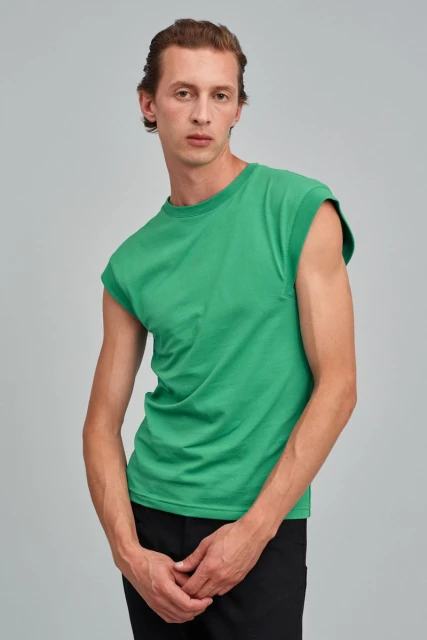 A fair-skinned adult male poses indoors wearing a green short-sleeved shirt and black pants, shot against a light gray backdrop.