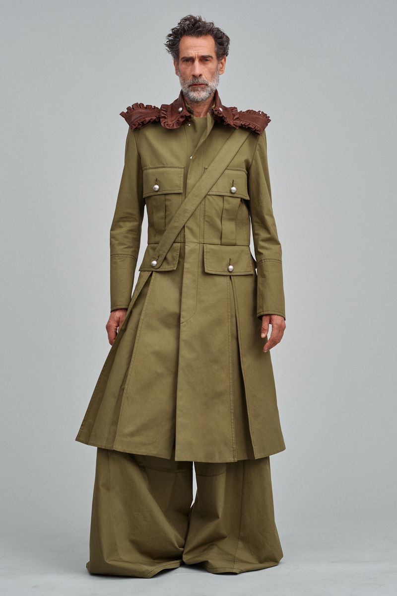 Full-length studio shot of a middle-aged male model with a grey beard wearing a green trench coat with wide leg pants on a grey backdrop.