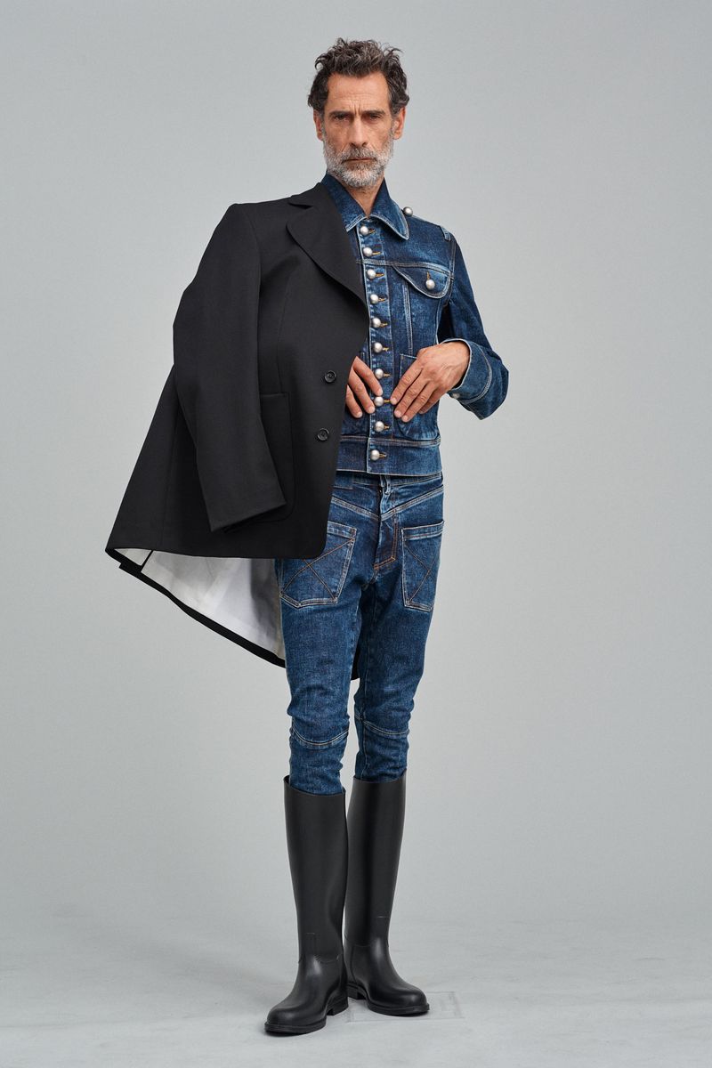 Man in denim jacket and jeans with a black coat posing in a studio, showcasing fashion-forward style with black boots and grey backdrop.