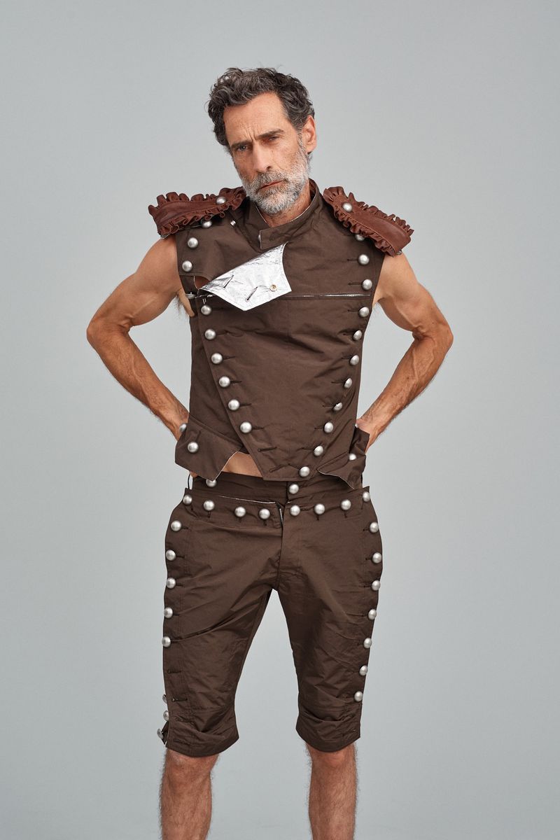 An adult male model poses in a brown vest and shorts with silver buttons in front of a gray background in a studio setting.