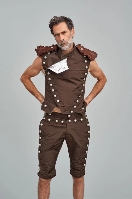 An adult male model poses in a brown vest and shorts with silver buttons in front of a gray background in a studio setting.
