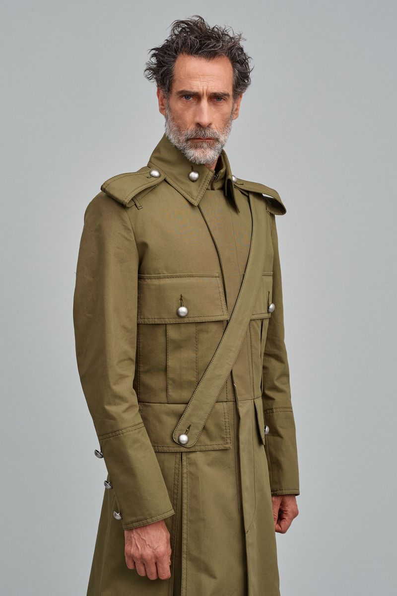 Mature man with a gray beard wearing an olive green military-style coat in a studio shot against a light gray background.
