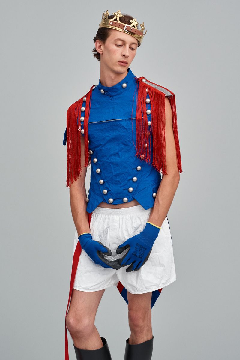 Male model poses in a royal-themed fashion outfit with a blue vest, red fringe, crown, white shorts, black boots, and blue gloves on a gray backdrop.
