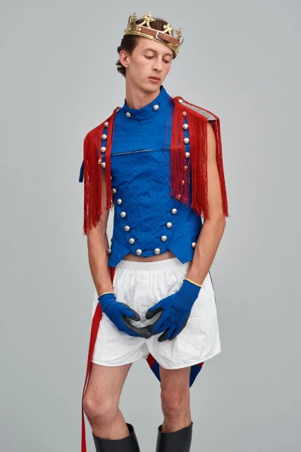 Male model poses in a royal-themed fashion outfit with a blue vest, red fringe, crown, white shorts, black boots, and blue gloves on a gray backdrop.