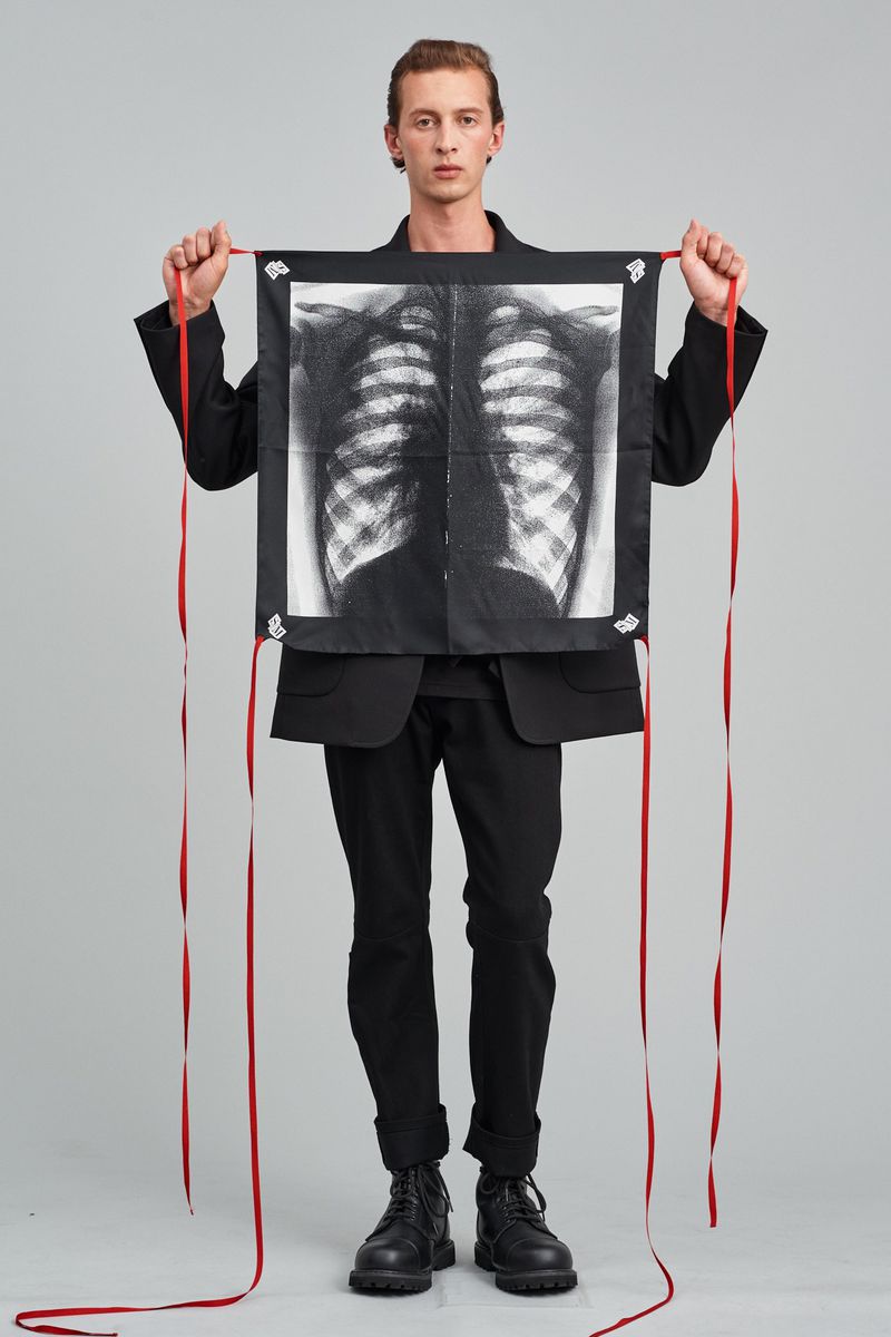 An adult male model in a black jacket holds a hanging square cloth with a ribcage x-ray design and red ribbons, in a studio setting.