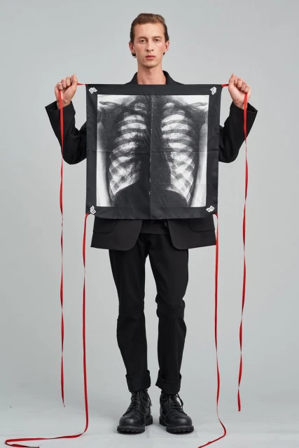 An adult male model in a black jacket holds a hanging square cloth with a ribcage x-ray design and red ribbons, in a studio setting.