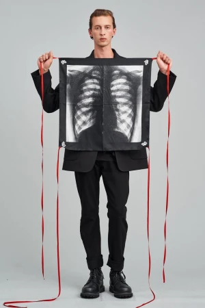 An adult male model in a black jacket holds a hanging square cloth with a ribcage x-ray design and red ribbons, in a studio setting.
