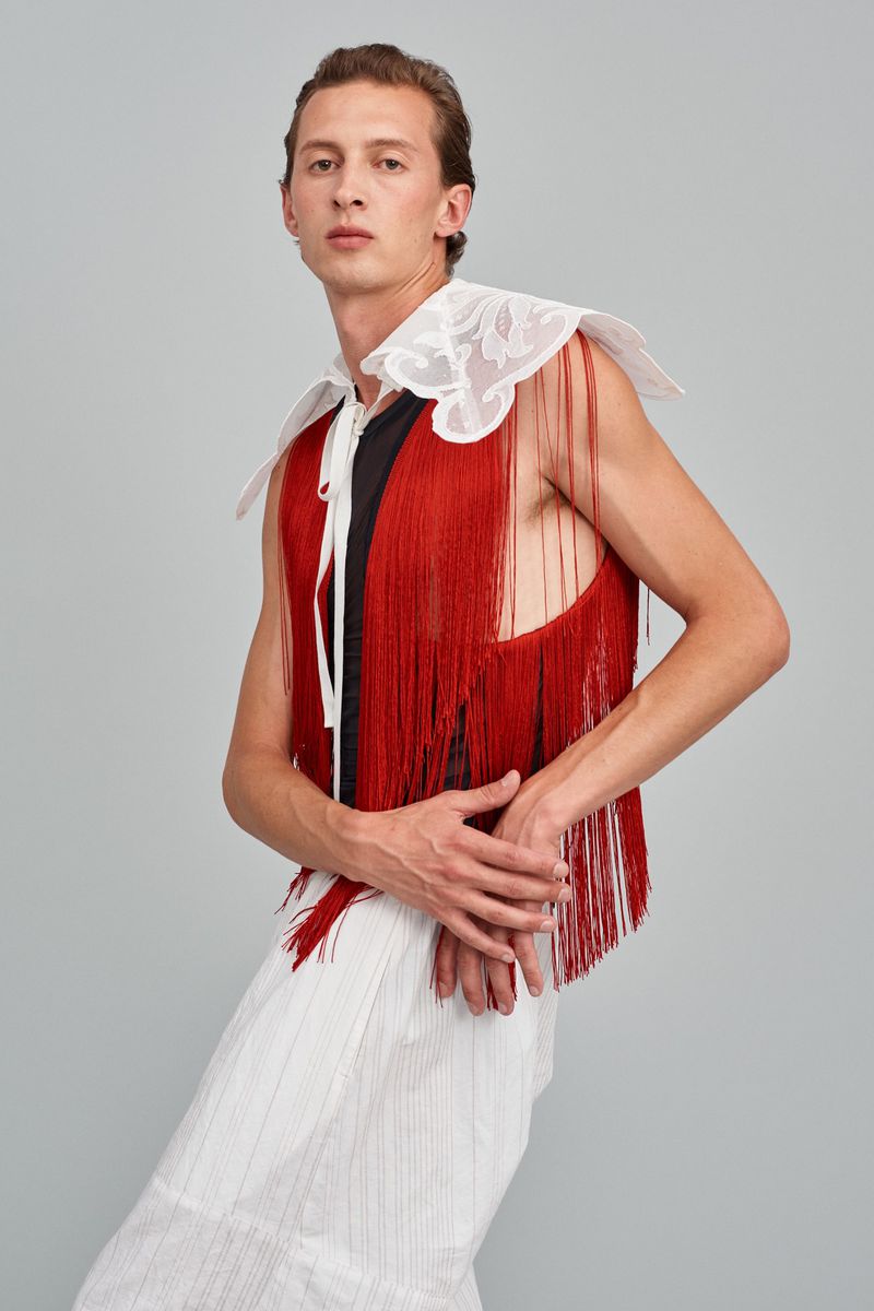 Androgynous male fashion model in sleeveless fringe top and pleated pants poses in a studio. Beauty and fashion editorial style.