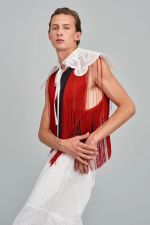 Androgynous male fashion model in sleeveless fringe top and pleated pants poses in a studio. Beauty and fashion editorial style.