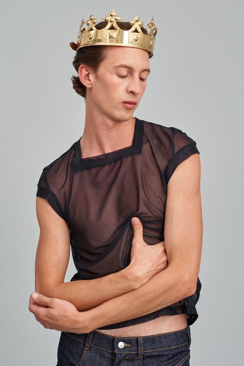 A slender man wears a gold crown and sheer black top, posing against a neutral backdrop in a fashion-forward studio portrait.