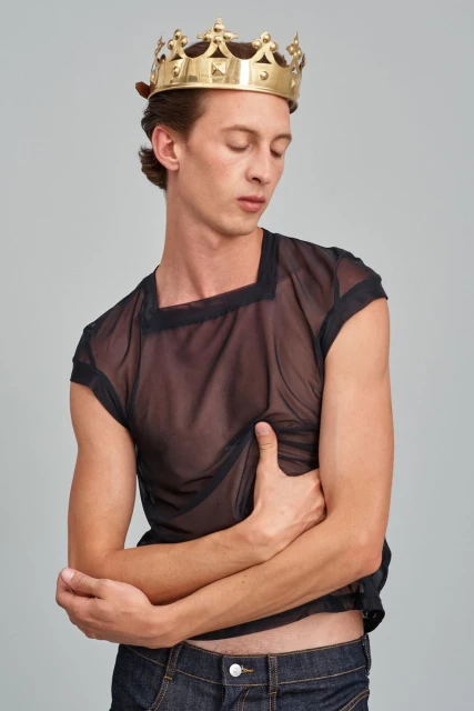 A slender man wears a gold crown and sheer black top, posing against a neutral backdrop in a fashion-forward studio portrait.