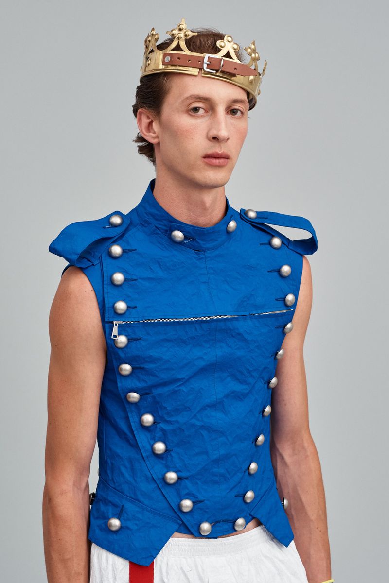 Male fashion model wearing a blue, button-up vest and a homemade gold crown posing indoors against a light-gray studio backdrop.