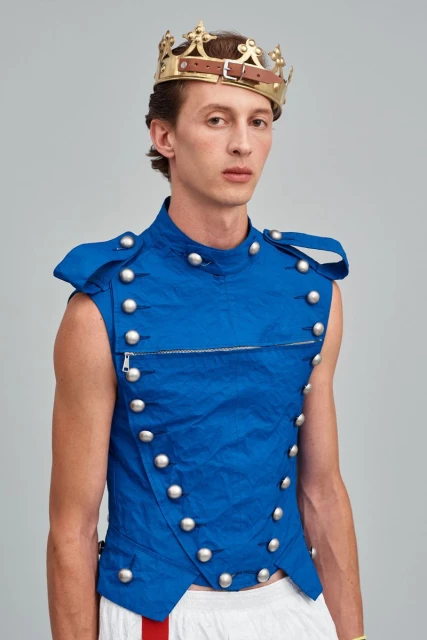 Male fashion model wearing a blue, button-up vest and a homemade gold crown posing indoors against a light-gray studio backdrop.