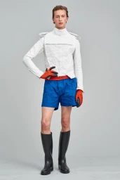 Model standing against a neutral grey backdrop wearing a textured white jacket, blue shorts, and tall black boots.