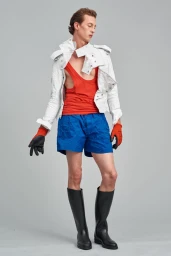 Male model in a white jacket, red tank top, blue shorts, and tall black boots against a grey studio backdrop